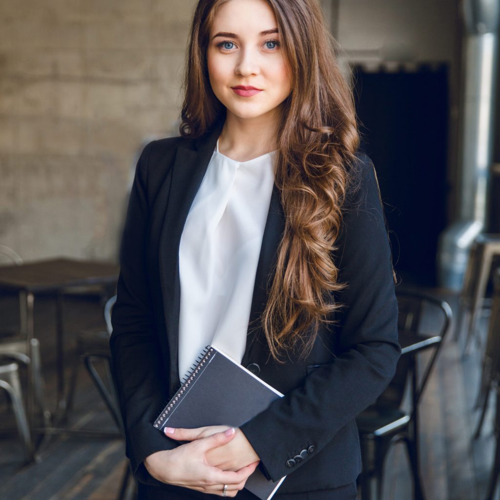 Brunette business woman with wavy long hair and blue eyes stands holding a notebook in hands. She wears a black suit and a white blouse. She is in a cafe with grey walls and dark furniture.